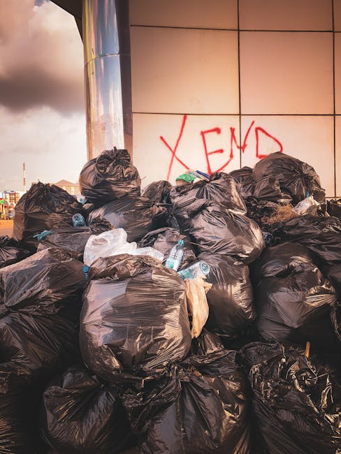 A person wearing a plaid shirt, grey trousers, and light green gloves is holding open a black rubbish bag prepared for waste disposal outdoors, with a background of green grass. The individual is posi