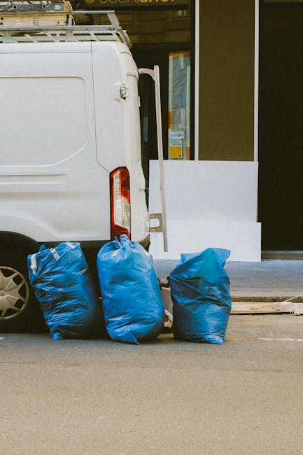 A large green wheeled rubbish bin filled with several red and black plastic bags labeled 'FLEXIBELDER CZE S1' and other text, positioned outdoors on a paved surface beside a dark brick wall. The bin a